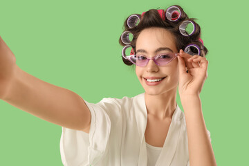 Young woman in robe with hair curlers and sunglasses taking selfie on green background, closeup