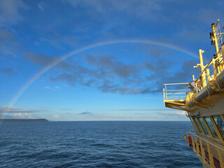 A vibrant rainbow arching over the blue ocean from the deck of a yellow ship under a partly cloudy...