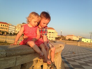 Two young siblings sit on a stone wall under the warm sun, sharing a quiet moment. Their...