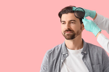 Doctor examining man with hair loss problem by using magnifier on pink background
