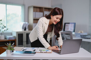 Young businesswoman leaning over her desk while working on a laptop and smiling, surrounded by...