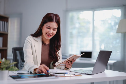 Smiling asian businesswoman holding notebook and analyzing marketing chart planning strategy, working with digital tablet and laptop computer at office, financial accounting concept