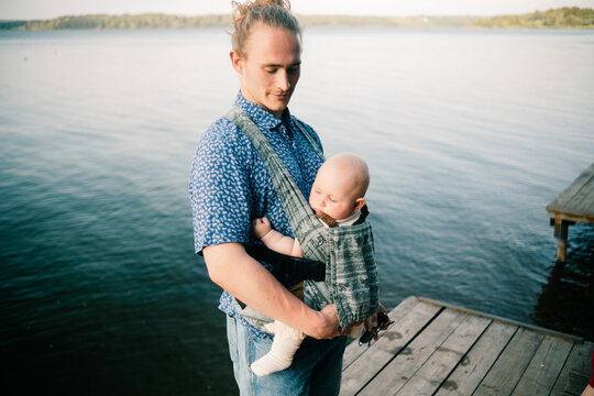 Portrait of young dad standing by lake holding six month old baby in sling loving outdoor family moment serene nature background