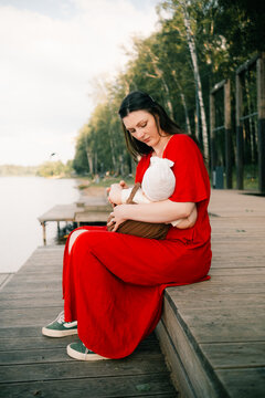 young mother in a red dress breastfeeding baby during summer walk near lake and forest serene natural outdoor family nurturing moment