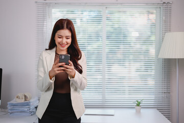 Young Asian businesswoman smiling while using her smartphone, standing in a modern office filled with natural light, surrounded by a desk, computer, and organized paperwork