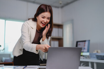 Asian businesswoman is standing at her desk, laughing and pointing at her laptop, enjoying a video call or celebrating a success in her modern office