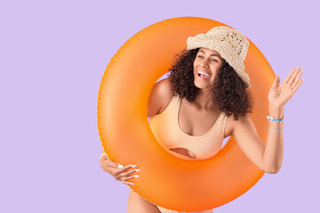 Young African-American woman in swimsuit with orange inflatable ring waving hand on lilac background