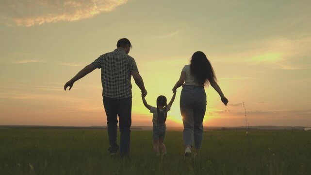 A happy family runs across the field with a small child at sunset in the sky, running mom, dad and kid are jumping cheerfully in the field in the evening, the team is traveling cheerfully playing.