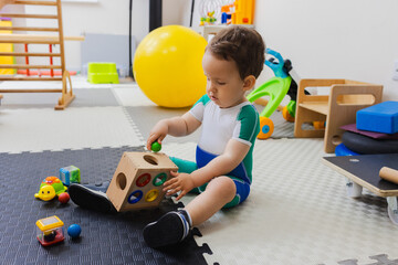 Obraz na płótnie Canvas Cute boy in compression suit during play therapy in rehabilitation center with sorter in hands training attention and fine motor skills. Paediatric physiotherapy