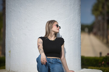 Young, beautiful woman, blonde, wearing a black t-shirt, jeans, and sunglasses, sitting on white...