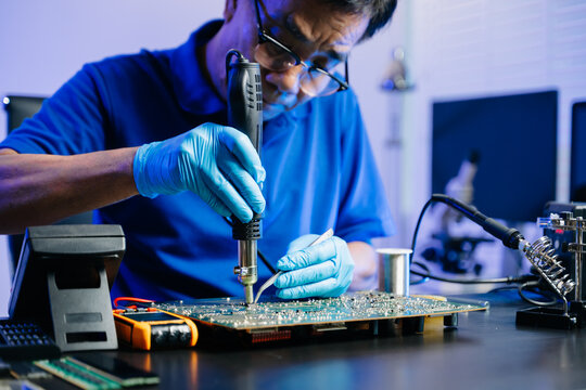 Senior technician soldering a circuit board on workbench. Blue gloves, precision tools, and electronics gear highlight real people
