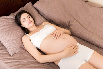 Young pretty pregnant woman, in pain, sitting on bed in bedroom, preparing to give a birth
