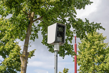 A modern white surveillance speed camera (flitspaal) mounted on a pole amidst green trees and a bright sky, ensuring safety and monitoring.
