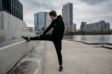 Man stretching leg on concrete wall by river with modern city buildings in the background. Outdoor...