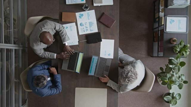 Full top view of three diverse male and female employees in suits working on business presentation at desk in office, using laptop computers, examining data charts and graphs