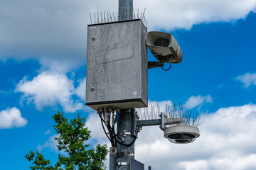 A concrete surveillance box sits on a tall pole alongside a camera and speakers. Spiky deterrents...
