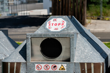 A metal waste disposal container stands prominently in an urban setting. Clear stop signs and...