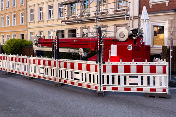 A large red construction vehicle is parked on a city street, surrounded by temporary barriers. The...