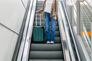 A young woman steps off the escalator at an airport, with only her legs and suitcase visible. The...