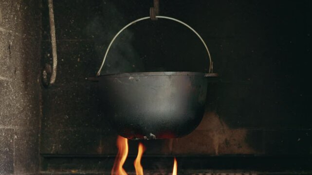Cast iron pot hanging over open fire in a rustic fireplace with smoke rising during traditional cooking.
