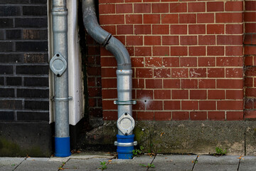 A metallic water pipe system is installed next to a red brick wall. The gray and blue pipes are...
