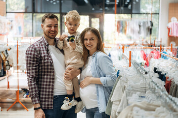 Standing and smiling. Father, son and pregnant mother are in kid's clothing store