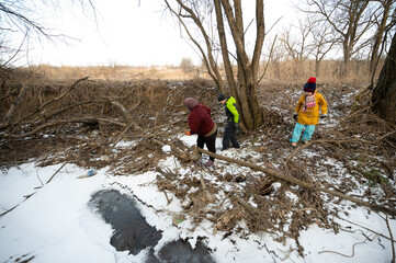 Children exploring a winter landscape while collecting sticks and playing near a frozen creek in a rural area during a sunny afternoon