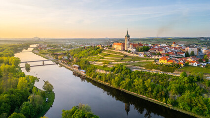 Fototapeta premium This aerial view captures Melnik City and its impressive chateau perched above the Labe River, surrounded by lush greenery and vibrant rooftops under a clear sky at sunset.
