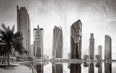 Monochrome city skyline with tall skyscrapers reflecting on calm water under a cloudy sky, Dubai...