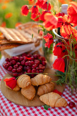 Close-up of fresh croissants, cherries, grissini, and poppy flowers on a picnic table with a red gingham tablecloth in a summer meadow.