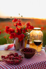 Close-up of summer lemonade with orange slices and mint in a glass jar, next to a glass of red wine and a bowl of cherries on a picnic table in a blooming poppy field.