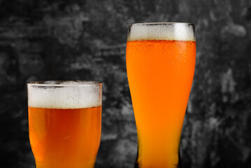 Two glasses of amber ale, one tall, one short, stand against a dark, textured background