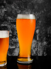 Two glasses of amber ale, one tall, one short, stand against a dark, textured background