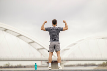 Rear view of strong sportsman standing on riverbank and flexing muscles.