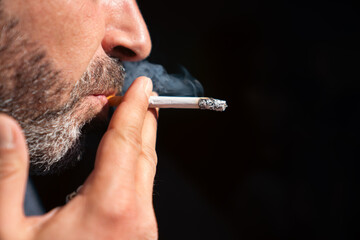 Close up of bearded man holding a lighter and smoking a cigarette on black background.