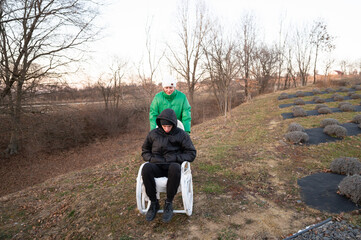 Two friends enjoying an adventurous moment in a wheelbarrow on a chilly afternoon in a quiet grassy area surrounded by trees and lavender fields