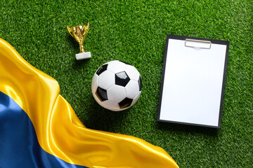 A soccer ball, golden trophy and clipboard with white paper sheet on a green field with a Ukrainian flag. High quality photo