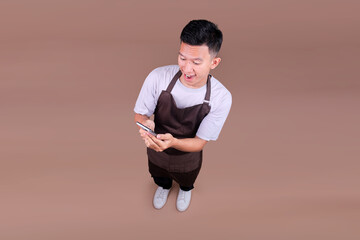Top View Of Excited Young Asian Male In Brown Apron Looking At His Phone