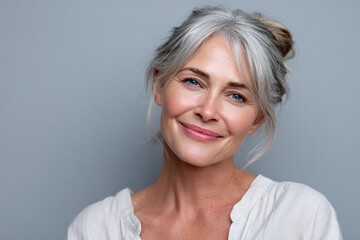 Fototapeta premium Beautiful mature woman with gray hair styled in a casual updo smiling gently at camera in a light-colored top against a soft gray background