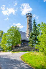 Fototapeta premium Svatobor lookout tower stands tall against a blue sky, surrounded by lush green trees. A well-maintained path leads to this popular tourist attraction in the serene countryside.