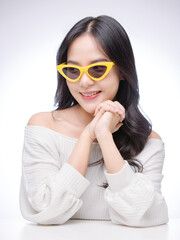 Asian young beautiful attractive cute happy woman long black hair poses with smile cheerful expression, wearing white sweater yellow sunglasses fashion, sitting white table against light background.