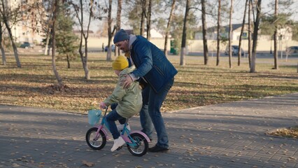father teaches child ride bike, girl pedals and wheels two-wheeled vehicle, fun horse game city...