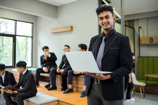 Portrait of Smiling Male Entrepreneur Holding a Laptop with Team in Background at Co-working Space