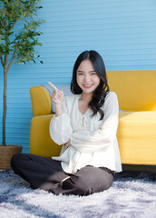Asian cheerful young beautiful happy funny woman wears casual clothes sitting on the floor in light blue living room at home  look at camera smiling posing points away finger for advertising content.