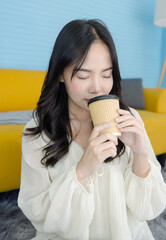 Asian young beautiful happy woman wears casual clothes sitting on the floor holding drinking coffee cup or hot beverage in light blue living room at home relax smelling morning coffee warming concept.