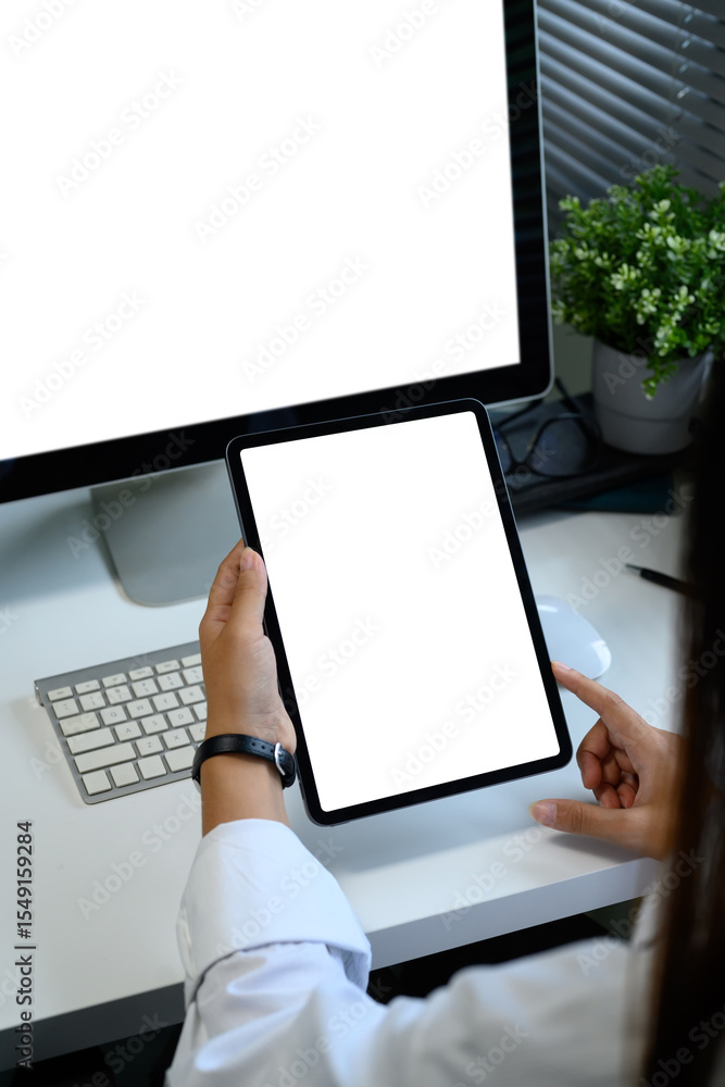 Poster closeup of hands holding a blank screen tablet in front of a desktop computer in a modern workspace.