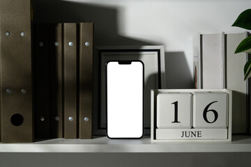 Smartphone with blank white screen standing on shelf next to calendar and books