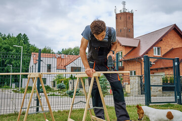 Man using electric jigsaw to cut wooden plank outside near residential buildings. Outdoor carpentry...