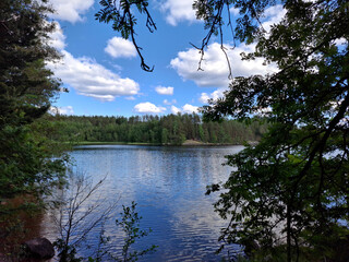 Still lake scene framed by dense forest and overhanging branches. Blue sky and clouds reflect off...