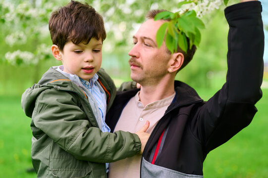 father and son enjoying nature on lush green park day. man holds child in arms under spring blossoms, showcasing warmth and family connection. perfect image for family-oriented themes